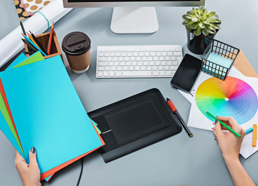 The desk with laptop, notepad with blank sheet, pot of flower, stylus and tablet for retouching. Workspace of professional retoucher. Creativity, design, art and modern equipments concept